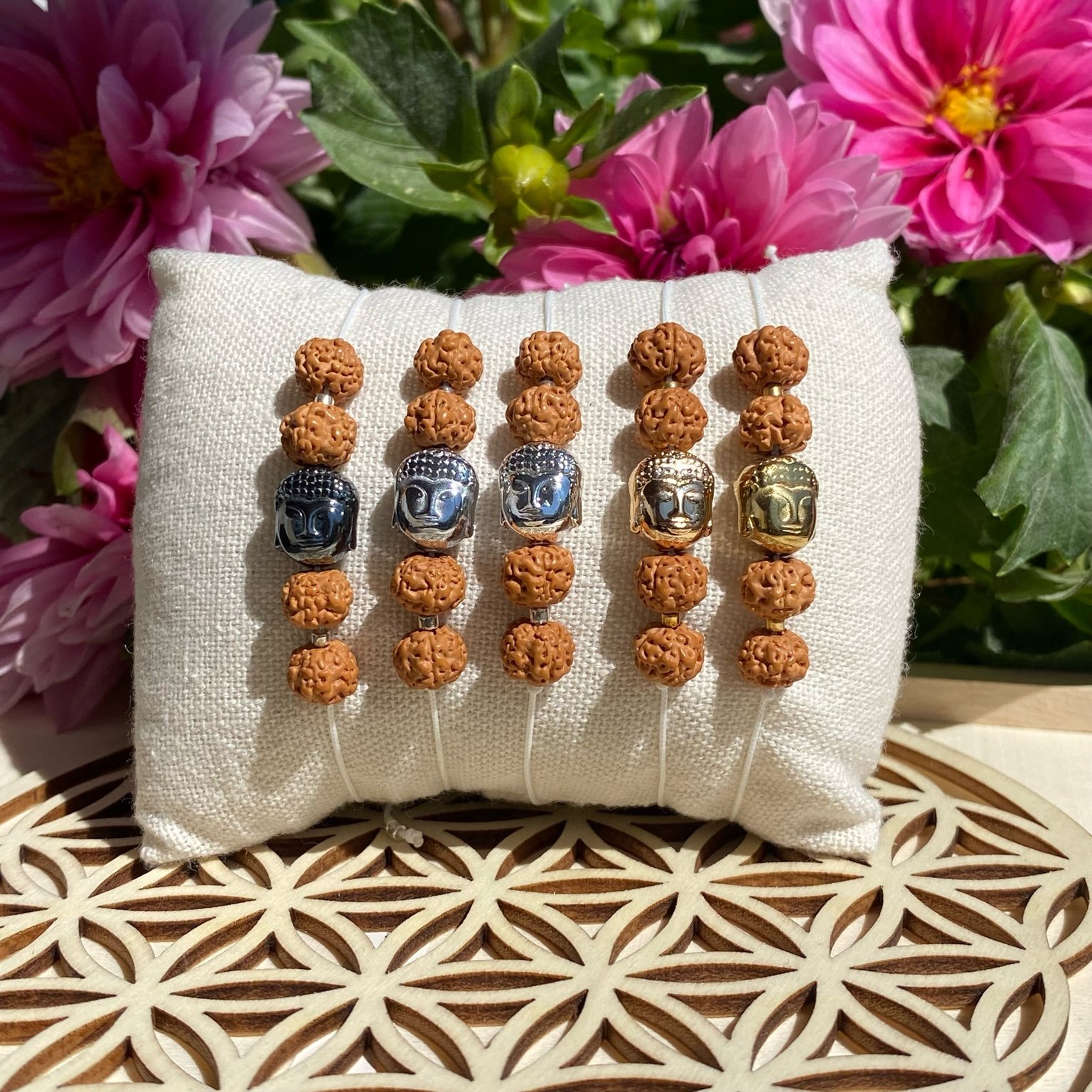 5 bracelets bohèmes en graines de Shiva avec perle tête de Bouddha en hématite, les 5 têtes ont des couleurs différentes allant du doré éclatant à l'argent foncé, fait main, présenté sur un coussin beige.
