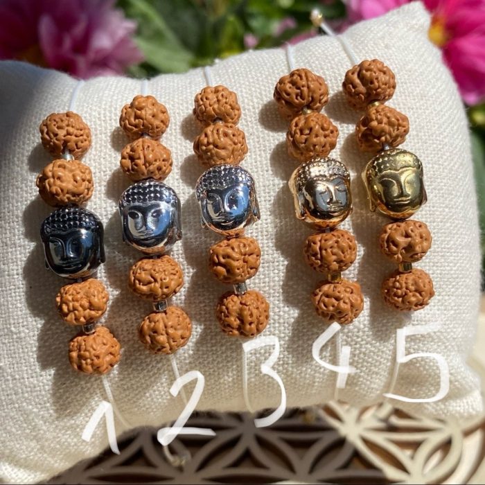 5 bracelets bohèmes en graines de Shiva avec perle tête de Bouddha en hématite, les 5 têtes ont des couleurs différentes allant du doré éclatant à l'argent foncé, fait main, présenté sur un coussin beige.