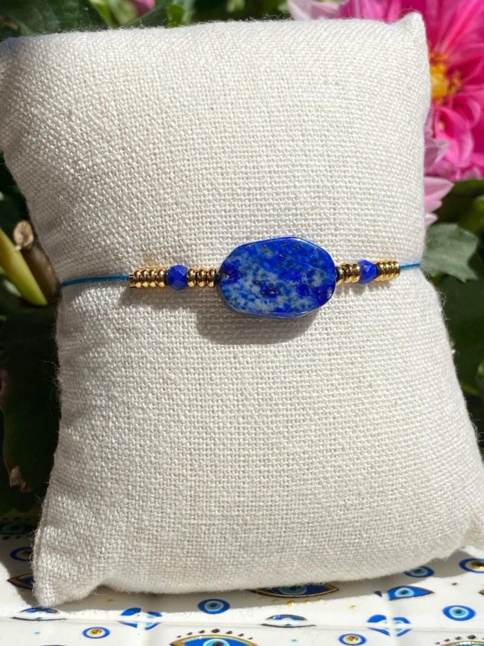 Bracelet bohème en lapis-lazuli (perle centrale rectangulaire et 2 petites perles facettées), avec des apprêts acier doré, posé sur un coussin beige