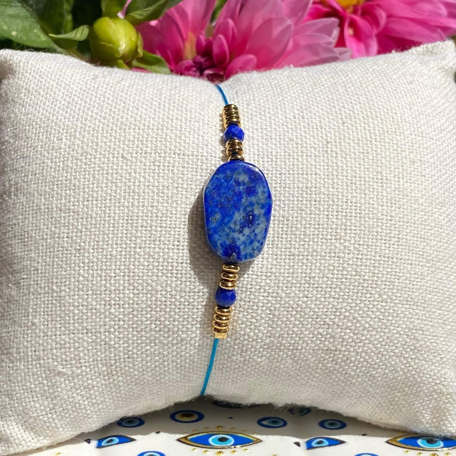 Bracelet bohème en lapis-lazuli (perle centrale rectangulaire et 2 petites perles facettées), avec des apprêts acier doré, posé sur un coussin beige