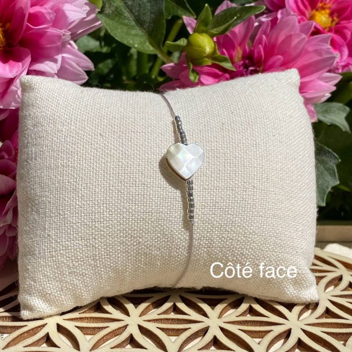 Bracelet bohème composé d'une perle centrale cœur en nacre, avec des perles argentées, posé sur un coussin beige