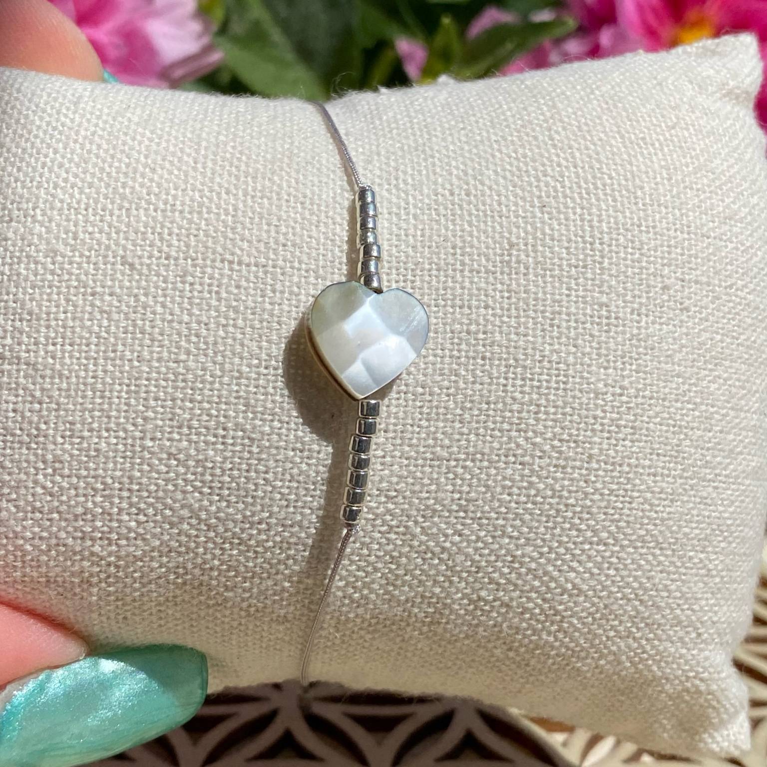 Bracelet bohème composé d'une perle centrale cœur en nacre, avec des perles argentées, posé sur un coussin beige