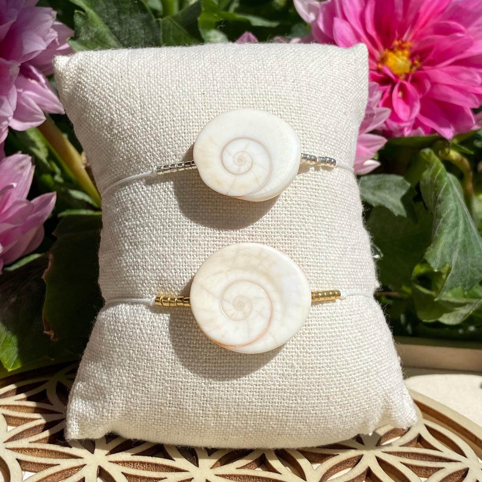2 bracelets œil de Shiva, un avec perles dorées et l'autre avec des perles argentées, posés sur un coussin beige