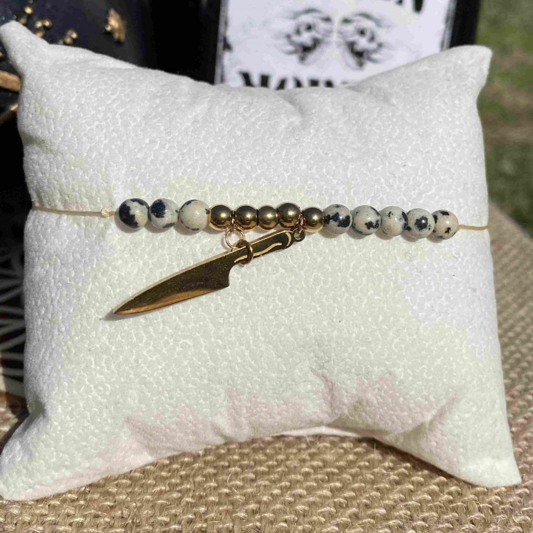 Bracelet inspirĂ© de Puglsey Addams, en jaspe dalmatien, apprĂȘts dorĂ©s en acier inoxydable et une breloque couteau dorĂ©. InspirĂ© par la sĂ©rie Netflix Wednesday et le film La famille Addams.